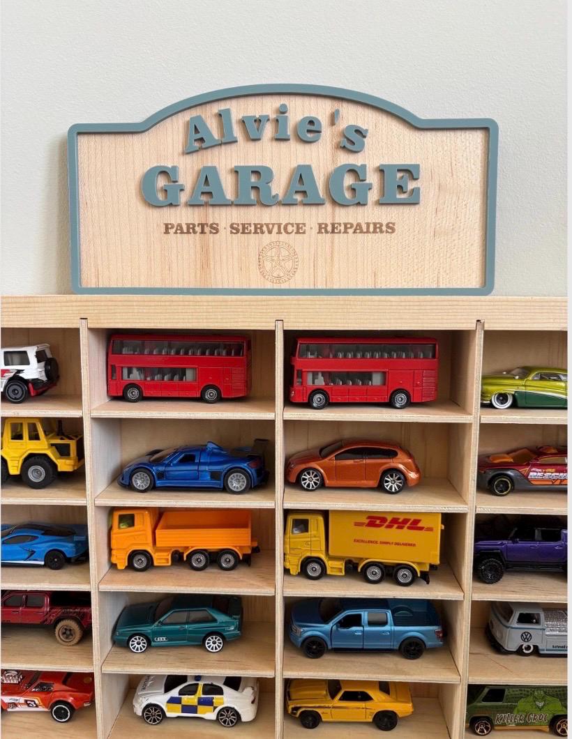 May include: A wooden display shelf with multiple compartments containing various toy vehicles, including buses, trucks, and cars. Above the shelf is a wooden sign with a blue border that reads Alvie's GARAGE PARTS SERVICE REPAIRS.